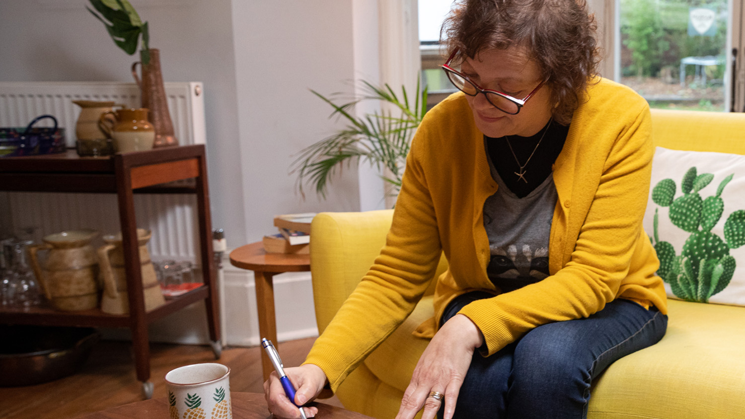 Woman wearing a yellow cardigan and glasses sitting on a sofa reaching forward to write on a document on a coffee table in front of her, with a house plant and window behind her.