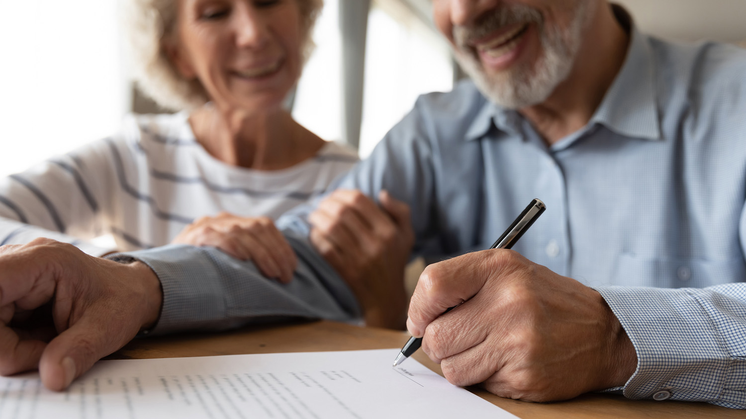An elderly couple sitting together, smiling warmly. The man, wearing a light blue checkered shirt, is signing a document with a black pen, while the woman, dressed in a white and grey striped top, rests her hand on his arm in a supportive gesture. The focus is on the man's hand and the document, with both appearing content and engaged in the process.
