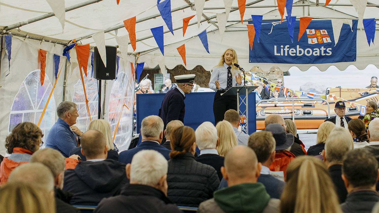 A group of people with their back to camera watching a female speaker on stage in a large gazebo with RNLI banners and bunting