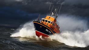 Yarmouth Severn class lifeboat is powering through the sea on a grey, stormy day with lots of white spray around it.