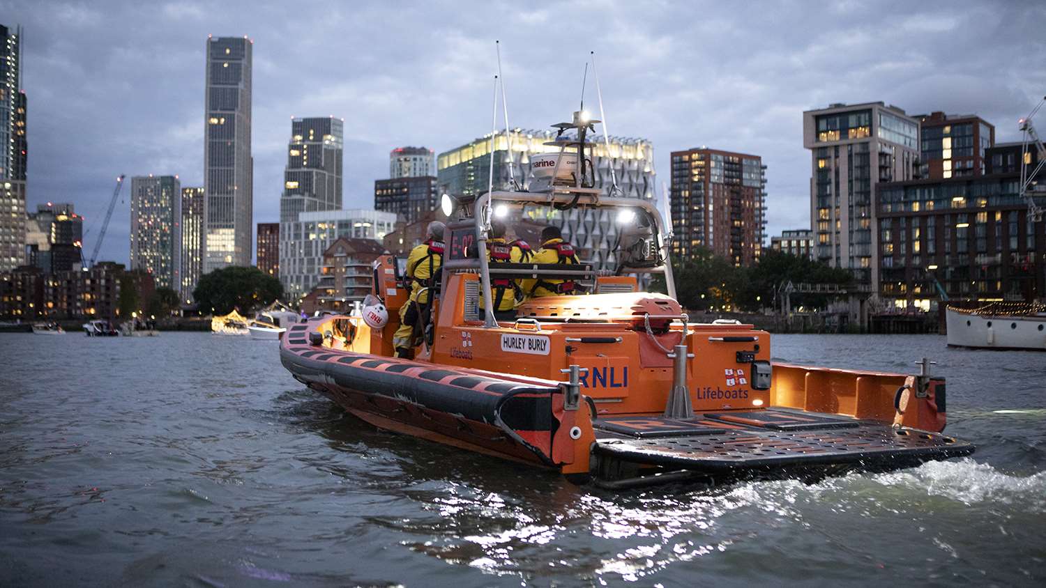 An E class lifeboat is on the River Thames in the evening, with office buildings and skyscrapers in the background.
