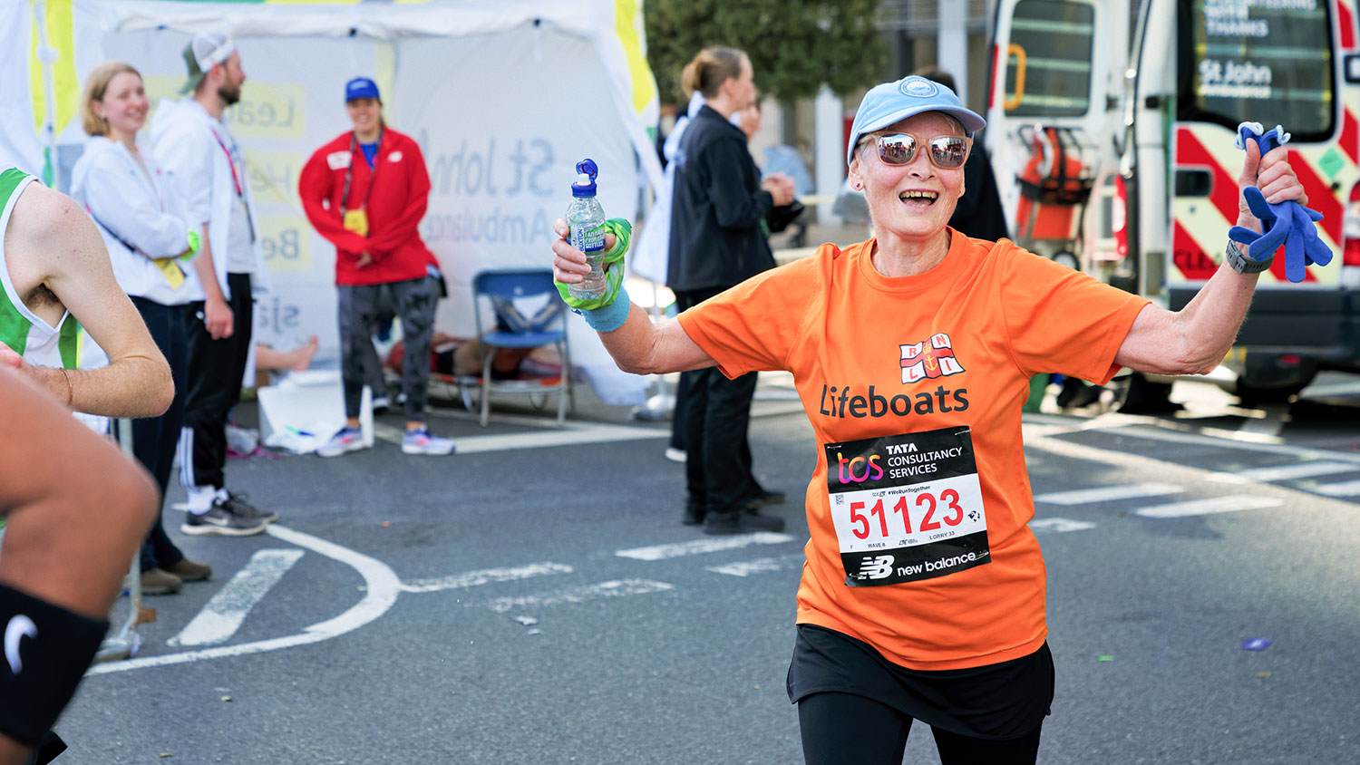 A smiling RNLI runner wearing sunglasses gives the thumbs up while passing a St John’s Ambulance tent during London Marathon