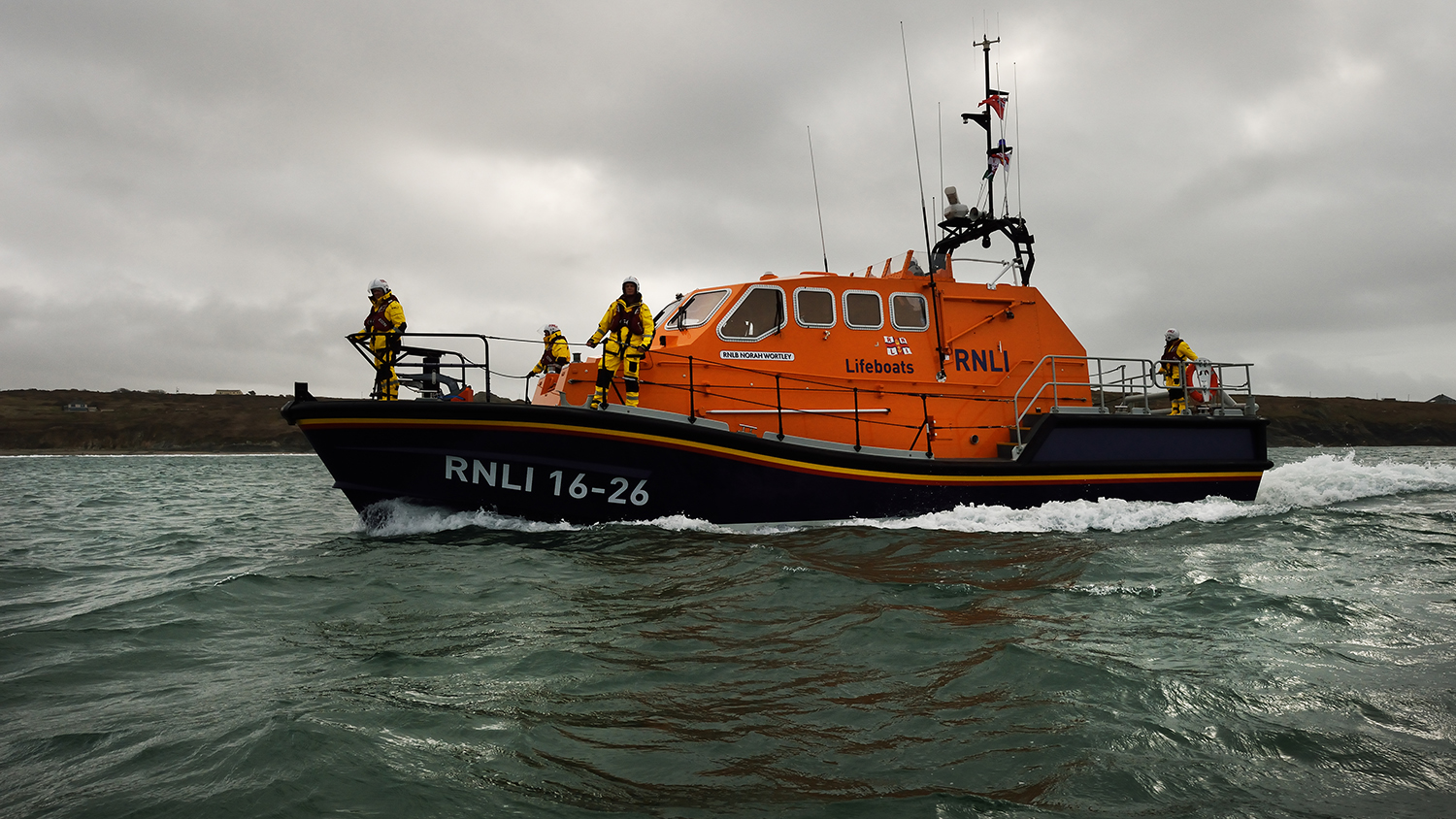 St Davids Tamar class lifeboat cuts a path through the sea, with four crew members wearing yellow all-weather kit standing on deck.