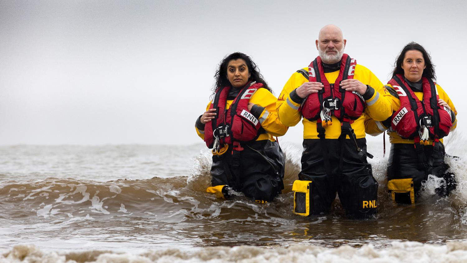 Three lifeboat crew members are stood waist-deep in the sea, looking at the camera with serious expressions on their faces.