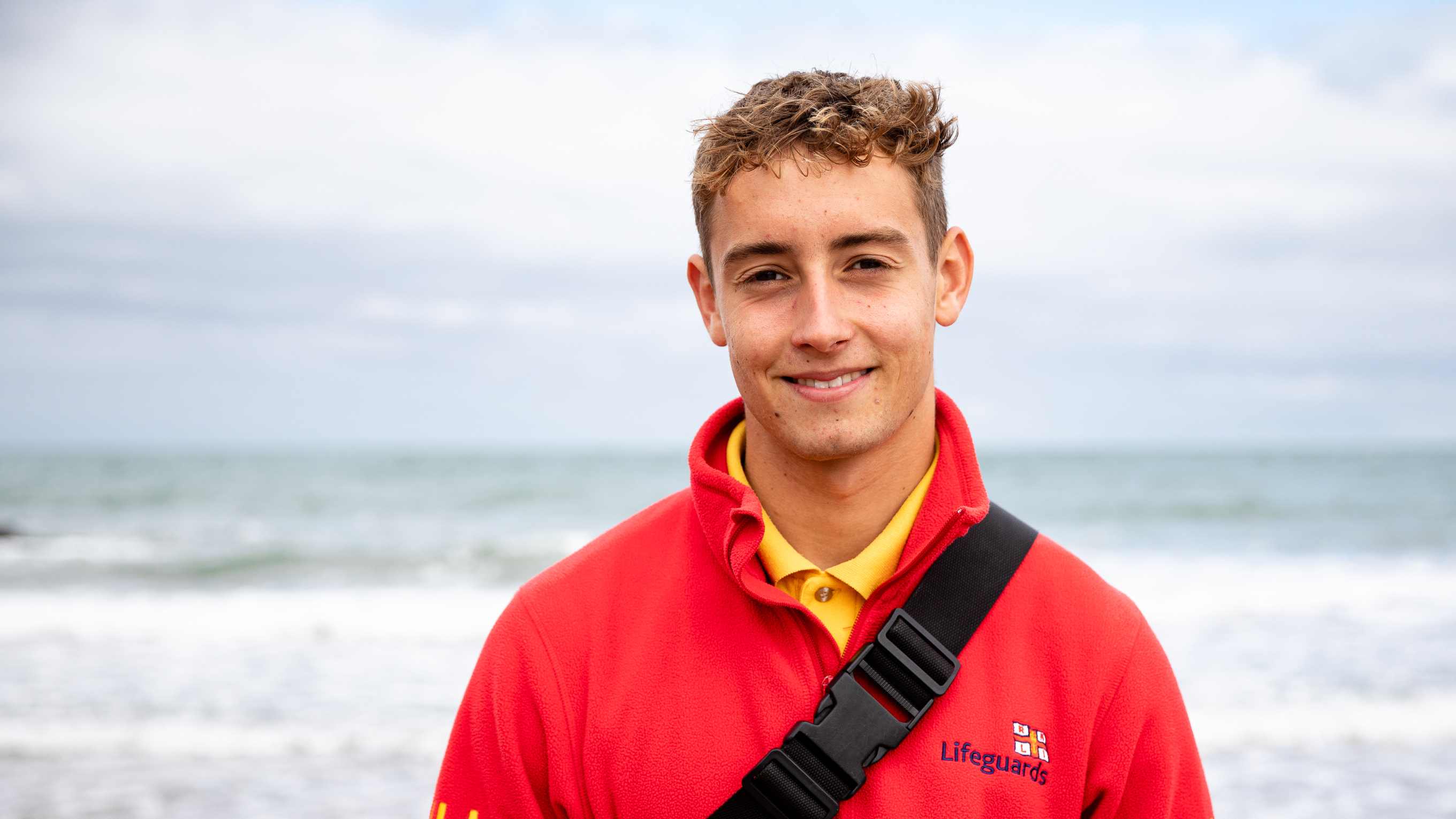 RNLI Lifeguard Hugo is smiling at the camera and is stood in front of the sea on Llangrannog Beach, Wales