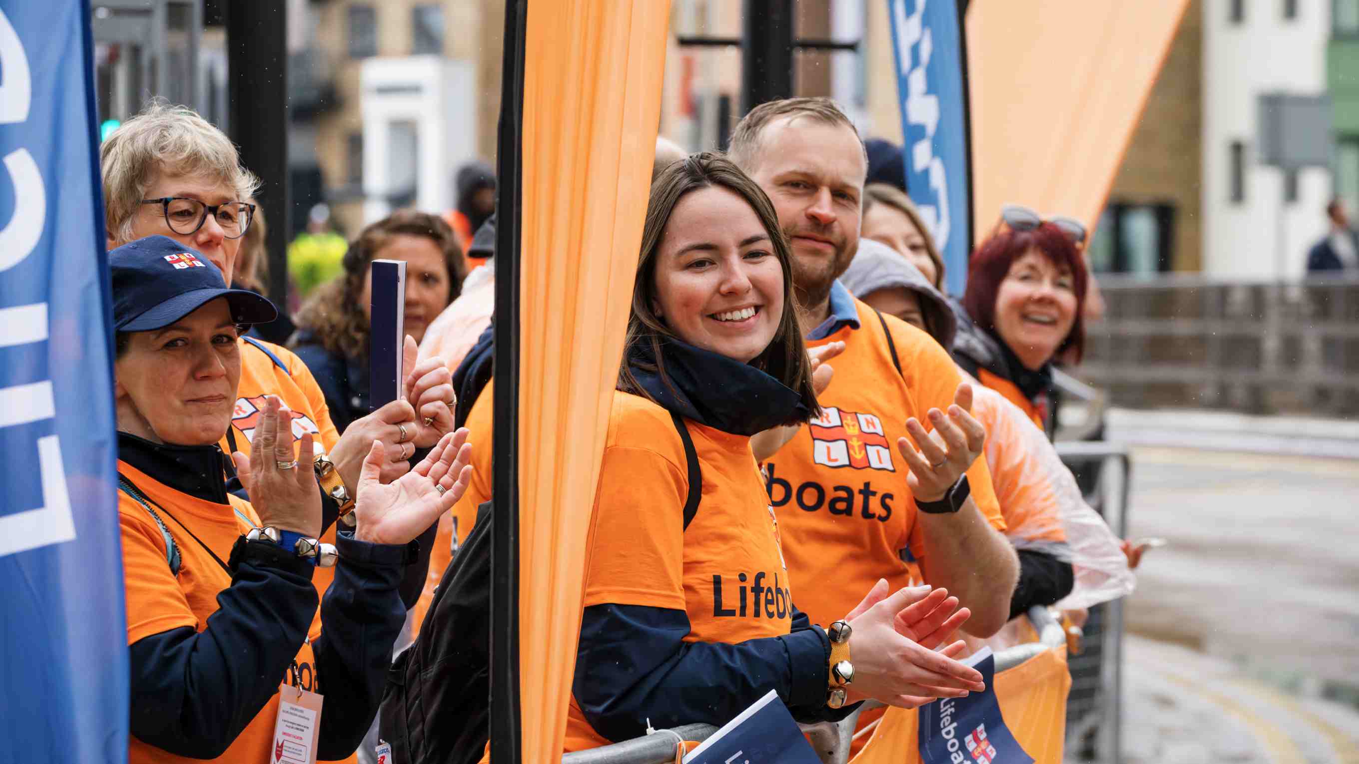 A group of men and women dressed in RNLI branded clothing are smiling and clapping their hands.
