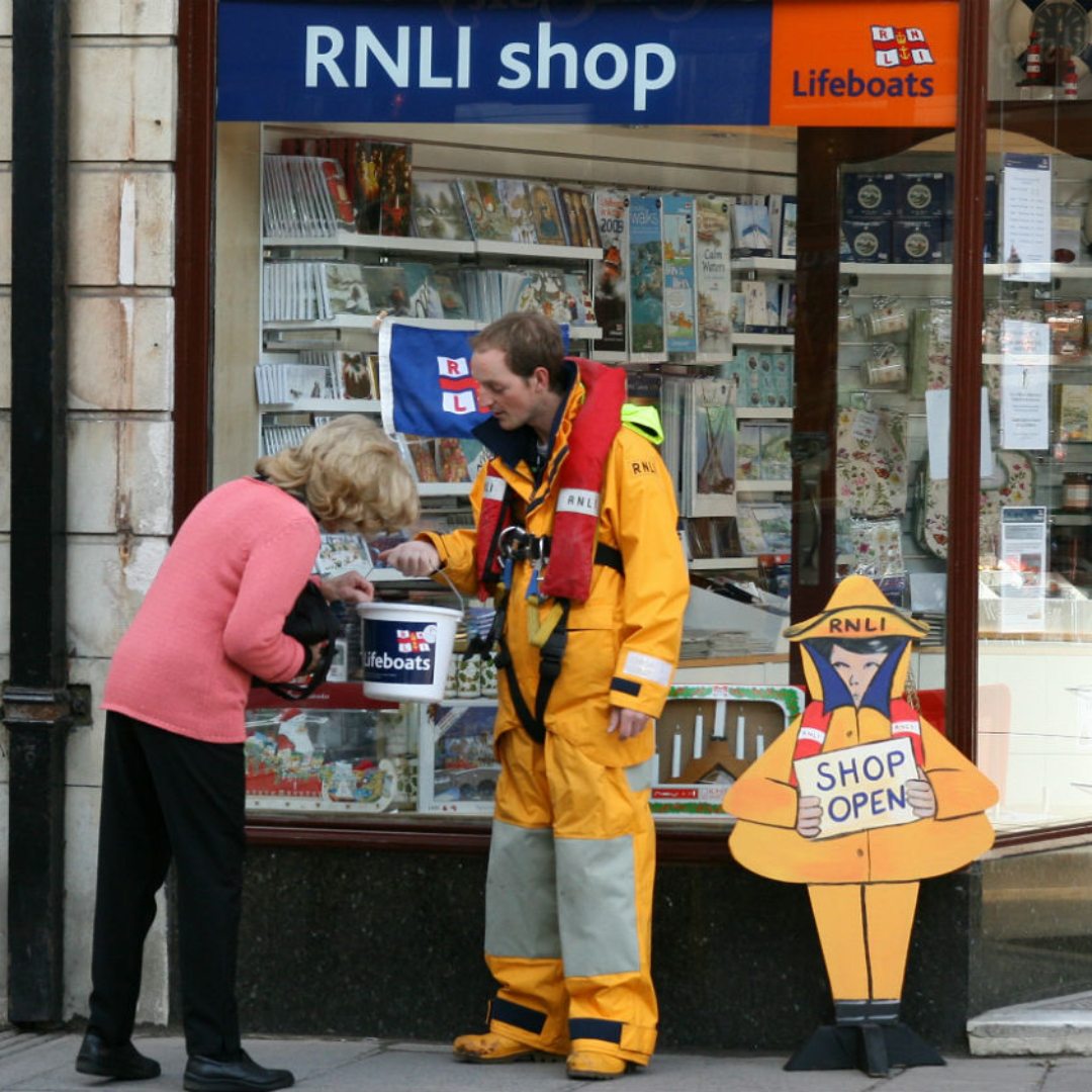 An RNLI fundraising volunteer in crew uniform collecting donations outside an RNLI shop in Swanage receiving a donation from a woman