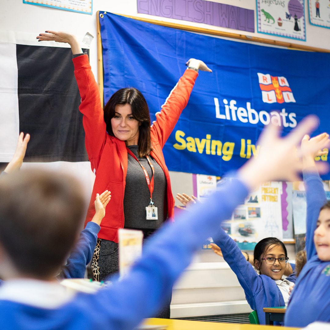 Water safety volunteer delivering a talk to schoolchildren in school, they all have their arms in the air