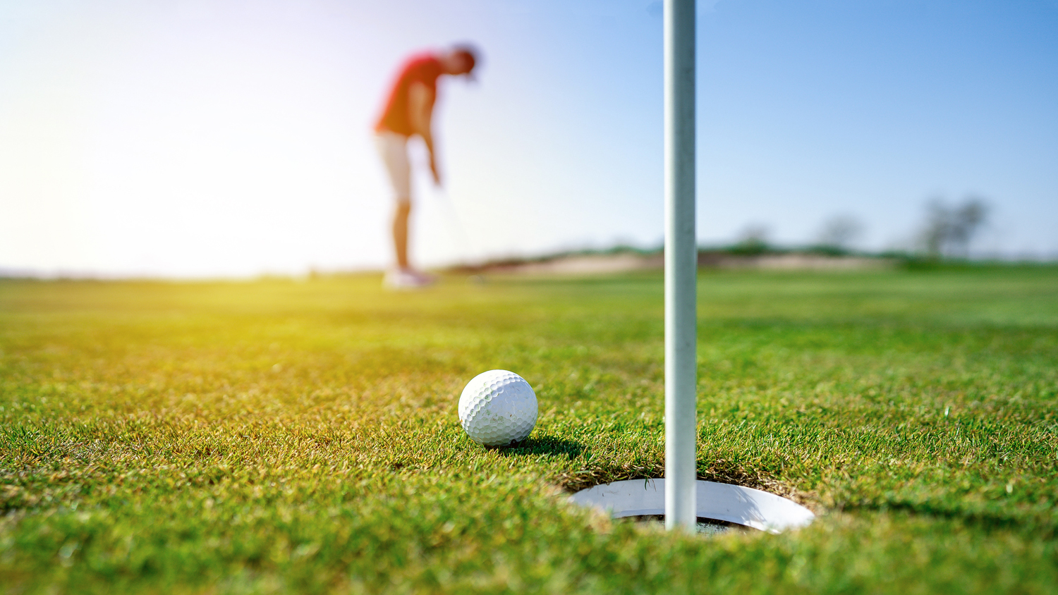 Close up of a golf ball on a green beside the hole with a golfer visible in the background