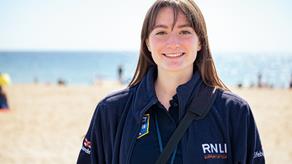A portrait of a young female RNLI staff member wearing a Respect The Water t-shirt and RNLI jacket. She is facing the camera and smiling while standing in front of a beach on a sunny day.