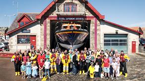 Group shot of volunteers including crew members, shore workers, fundraisers, families and children in front of Anstruther Lifeboat Station with Mersey class lifeboat Kingdom of Fife 12-17 on the slipway.
