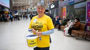 An RNLI fundraising volunteer is stood in a busy train station holding a branded bucket and smiling.