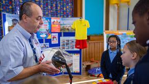 An RNLI water safety education volunteer is holding a lifeboat crew helmet and talking to a group of children in a school.