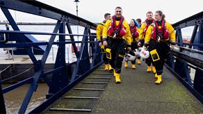 A group of lifeboat crew members wearing inshore lifeboat kit cross a bridge onto some docks.