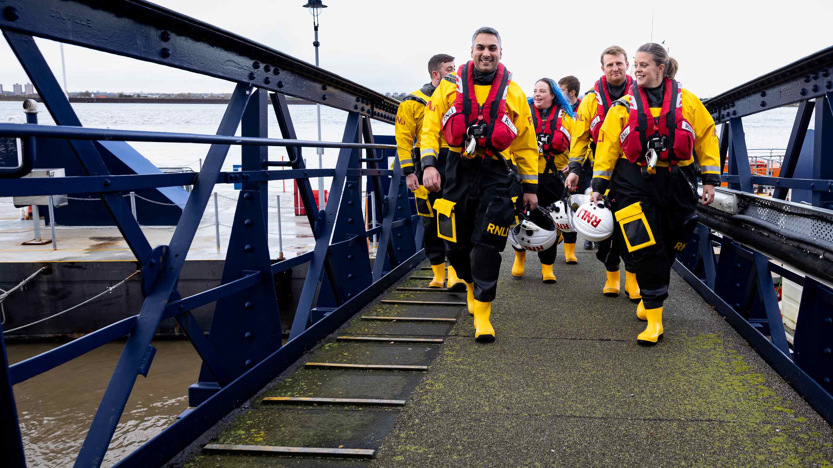 A group of lifeboat crew members wearing inshore lifeboat kit cross a bridge onto some docks.
