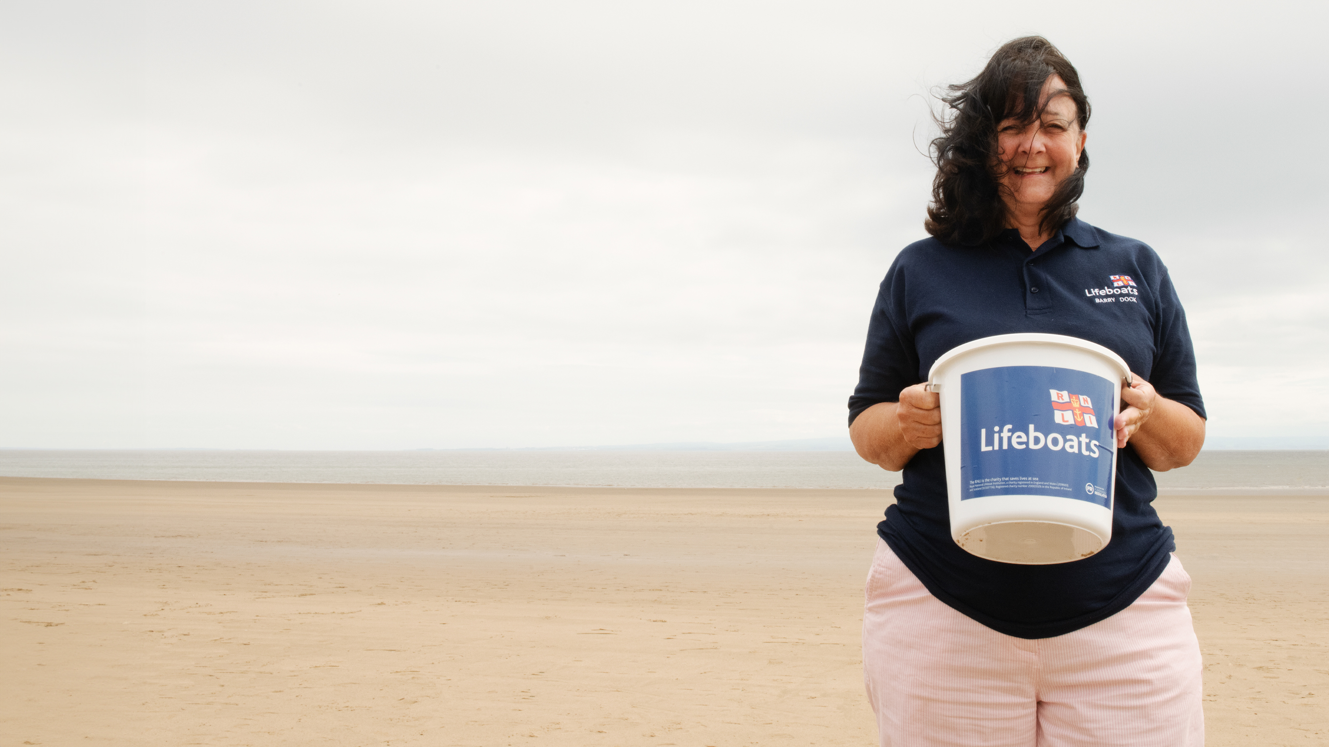 Barry Island Shop Volunteer Avril Kitchen stood on the beach holding a collection bucket.