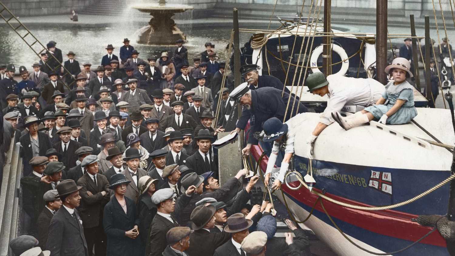 Celebrating the 100th anniversary of the RNLI, with a lifeboat in Trafalgar Square, London, surrounded by members of the public wearing hats