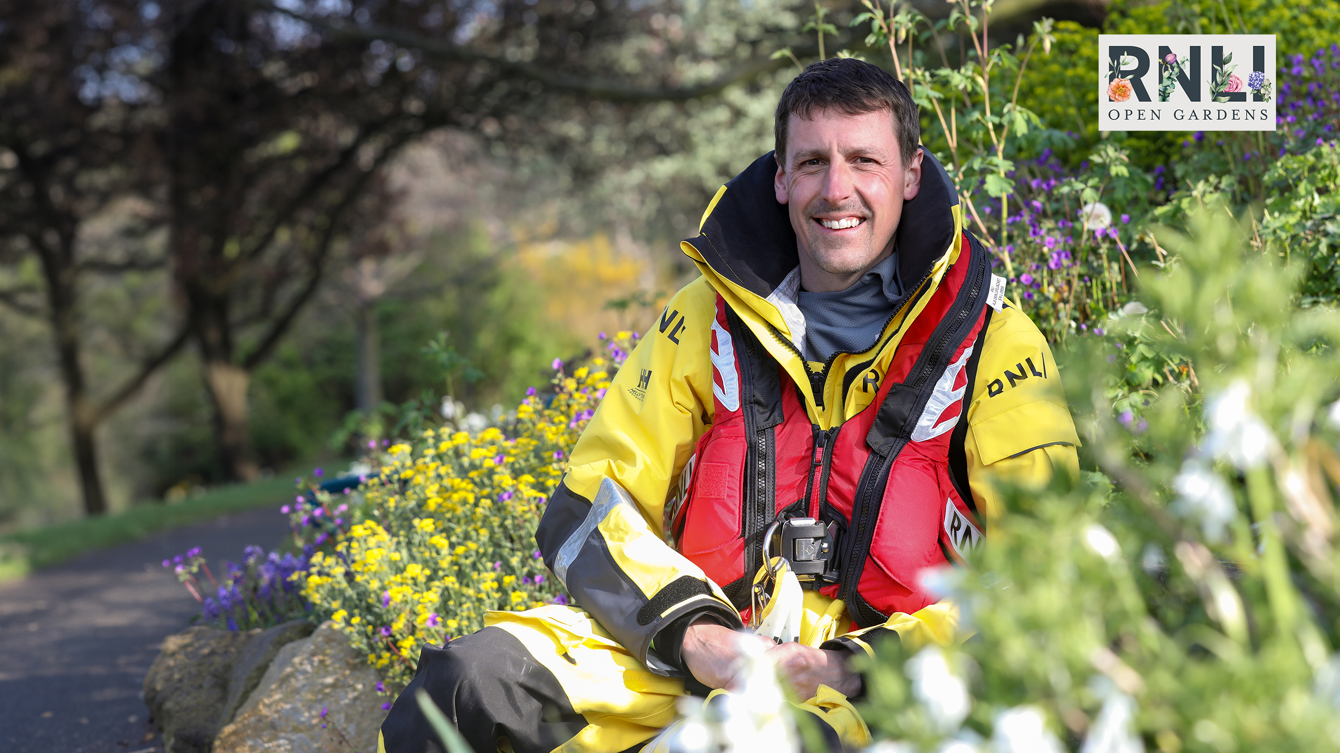 A white lifeboat crew member with short brown hair and a moustache sits in full kit among flowers, with trees in the background