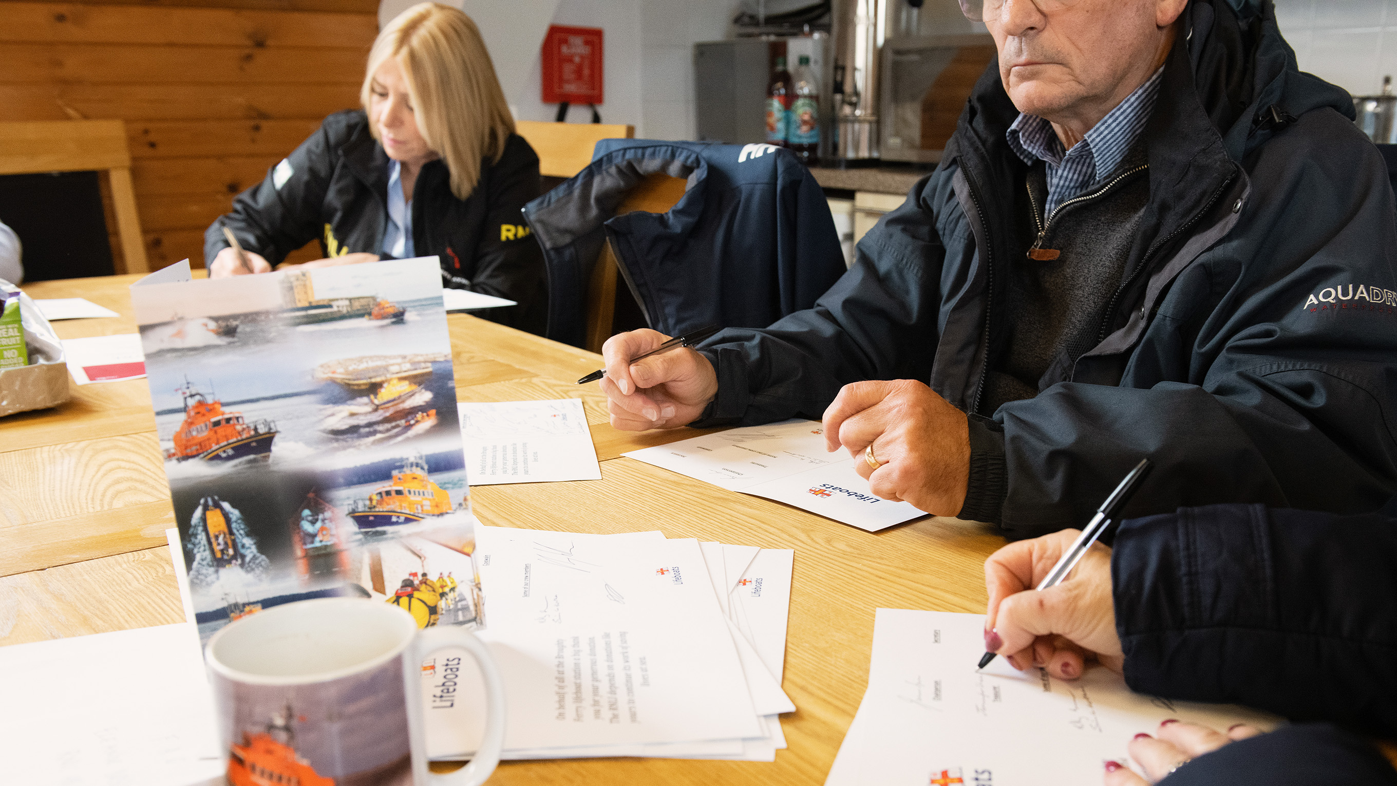 Volunteers gathered round a kitchen table writing things down on paper. On the table, there is a card with lifeboats on, as well as a mug with a lifeboat on.