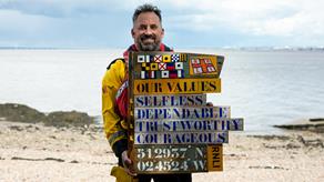 Portishead Helm, Andy Weston, is standing on a beach on a sunny day, holding a wooden board made up of the words of the RNLI values.