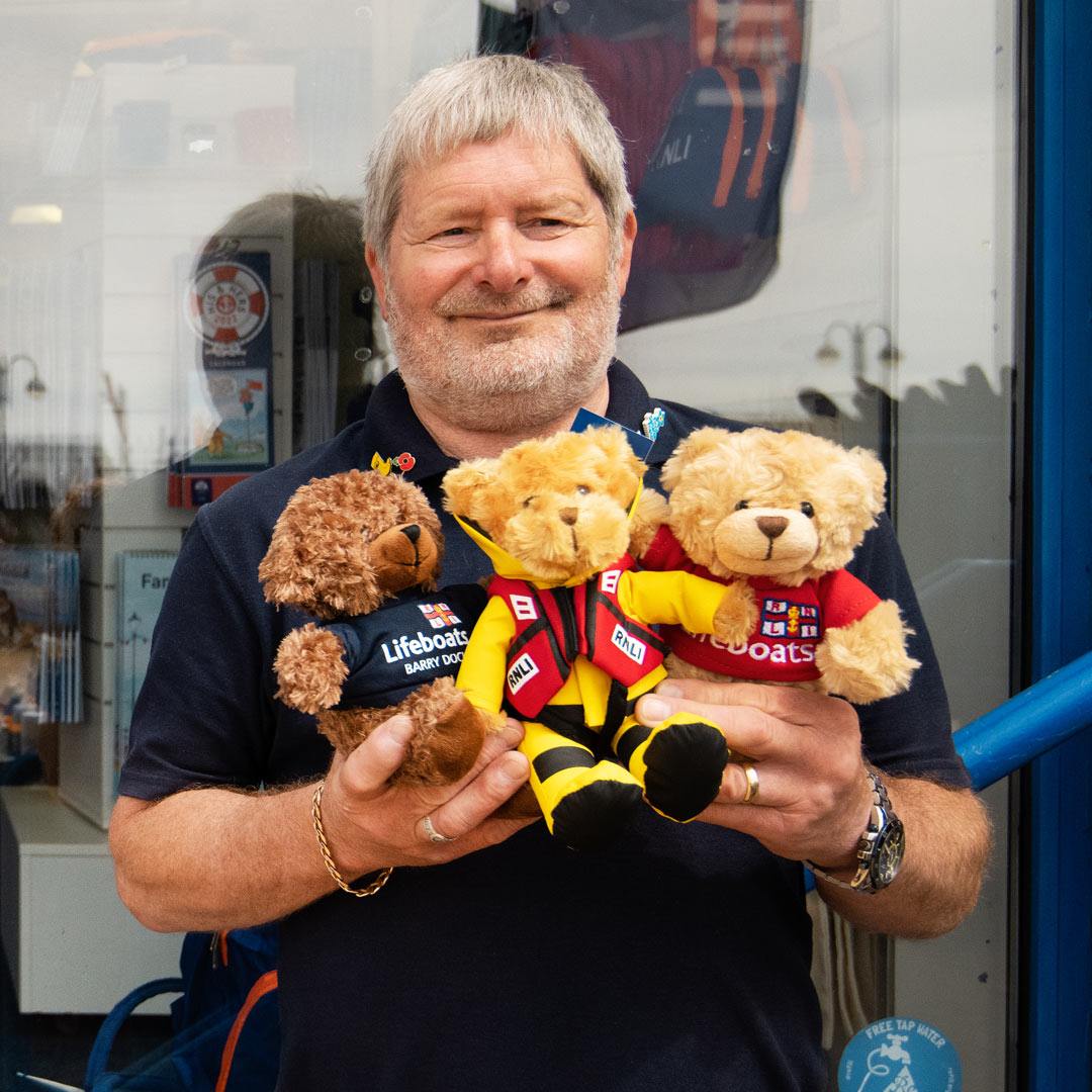 An RNLI shop volunteer stands outside of an RNLI shop holding a trio of RNLI teddy bears.