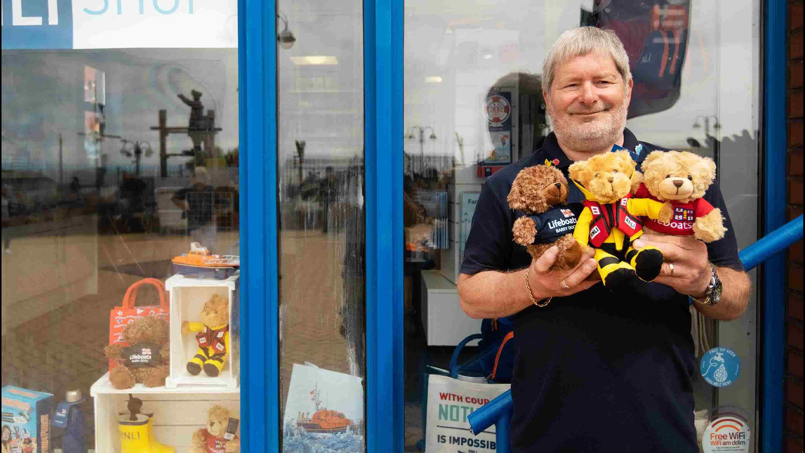 An RNLI shop volunteer stands outside of an RNLI shop holding a trio of RNLI teddy bears.