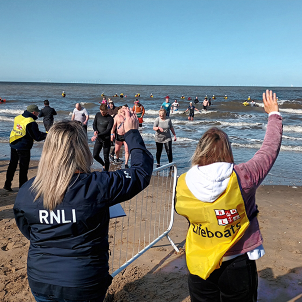 Two people wave at a group of swimmers heading out on a cold water dip