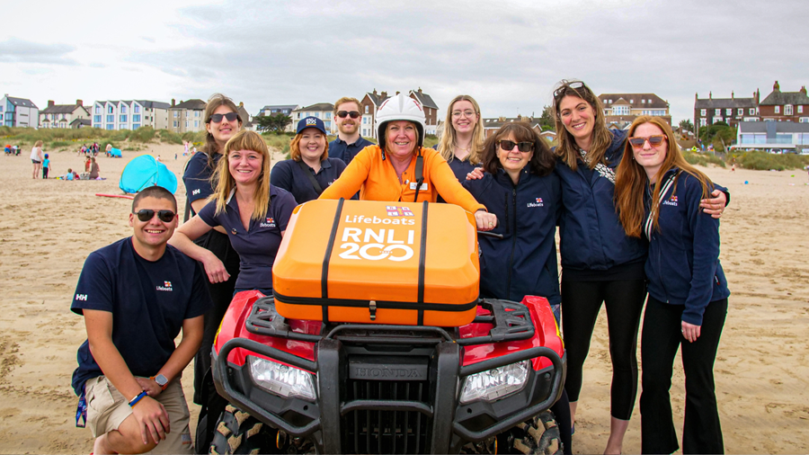 Group of people in RNLI clothing on the beach, smiling and gathered around a large orange case, perched on a quad bike