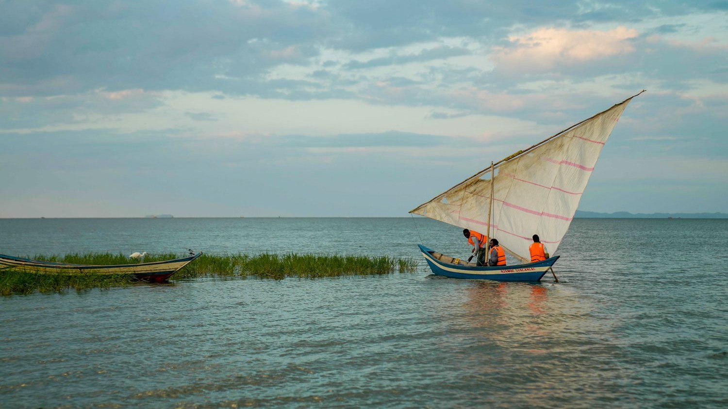 A landscape photo taken in Tanzania – in the foreground there are small buildings on green land, then in the background there a body of water with small boats on it.