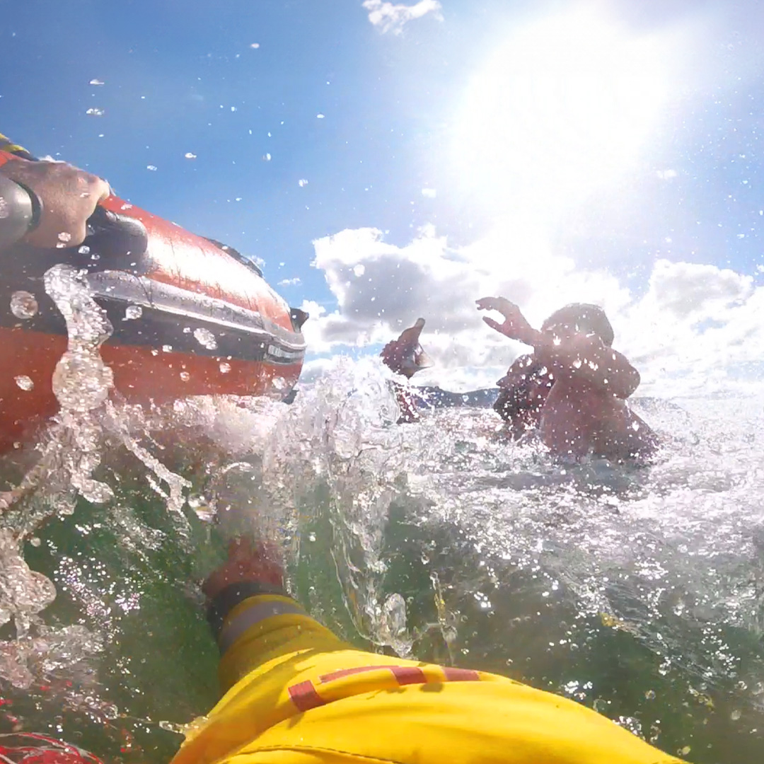 A screenshot from an RNLI lifeboat crew member’s helmet camera footage of a rescue. The crew member is in the water next to the lifeboat. A man is holding a child and they are in deep water, waving their arms.