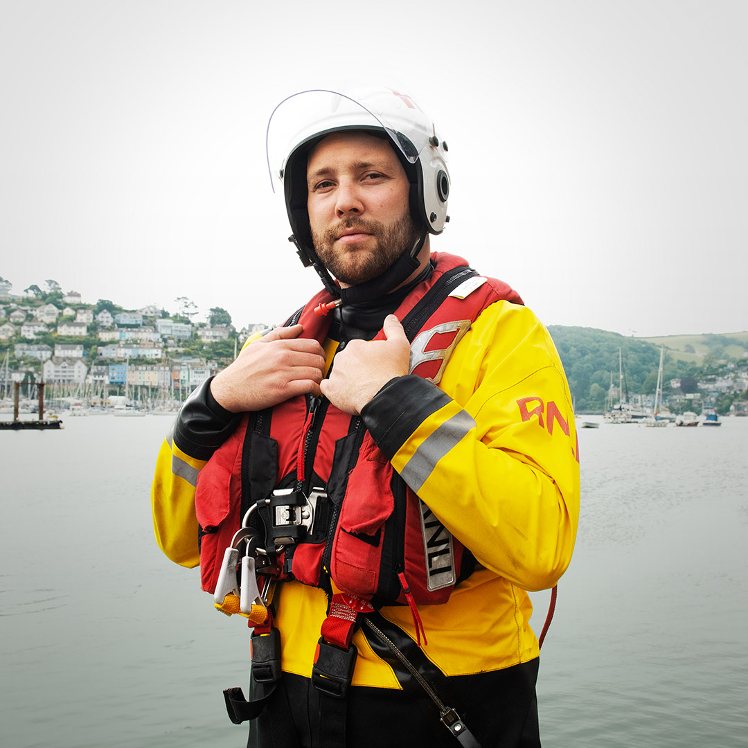 A male crew member, wearing his crew kit and a white helmet, looking directly at the camera. Behind him, you can see the coastal town of Dartmouth.