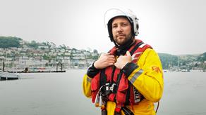 A crew member, wearing his crew kit and a white helmet, looking directly at the camera. Behind him, you can see the coastal town of Dartmouth.