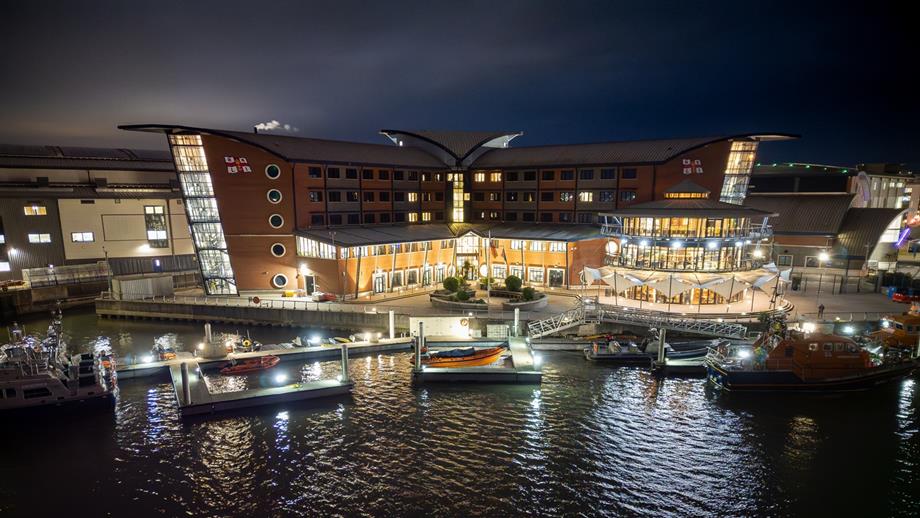 An aerial drone shot over the water of the RNLI College at night. It is lit up and lifeboats are moored at the college pontoon.