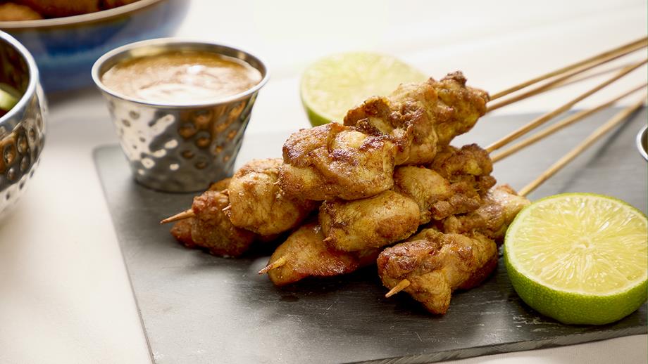 Chicken satay skewers alongside two lime halves and a pot of dipping sauce on a black slate serving dish.