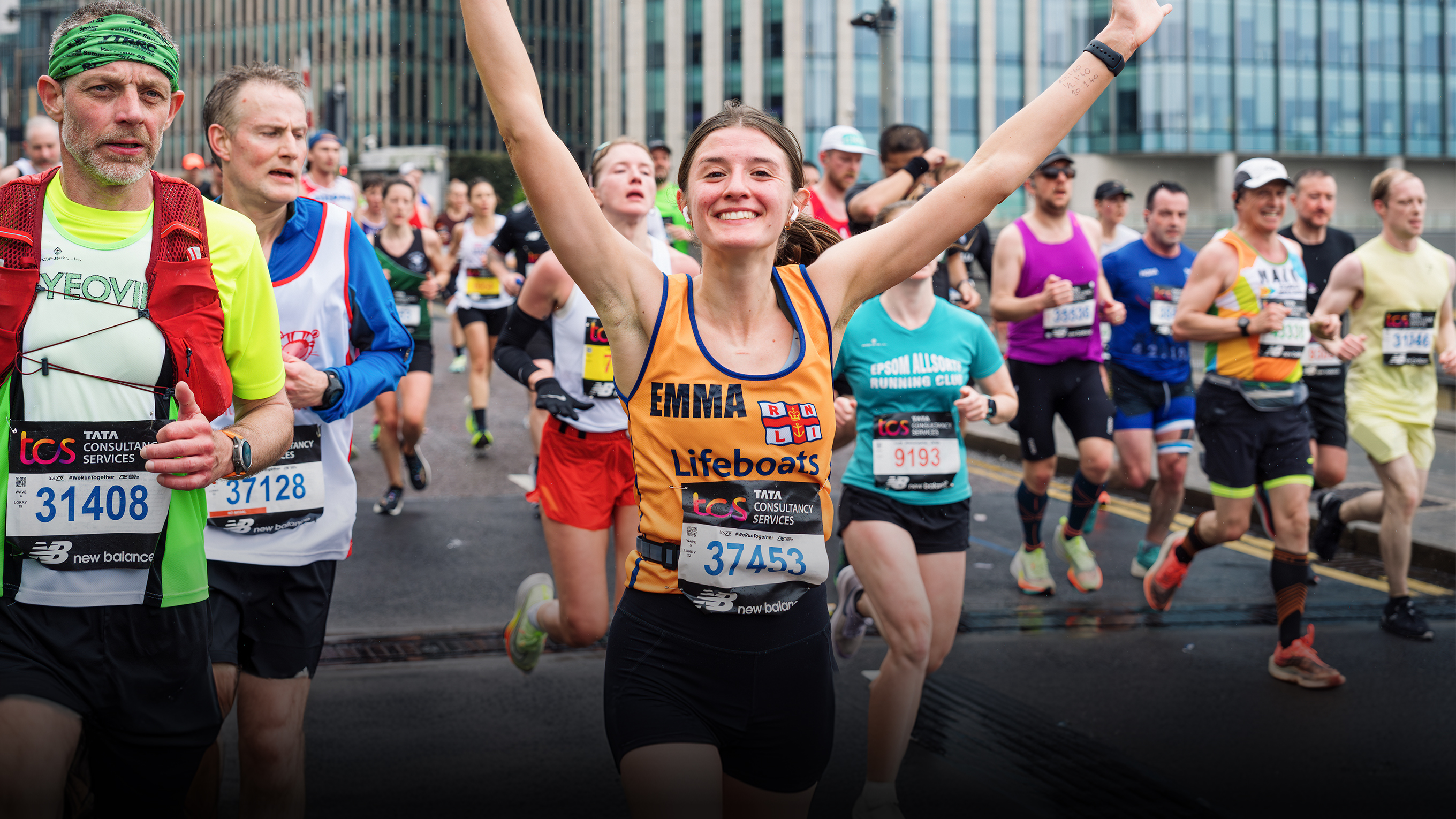 A woman running the London Marathon for the RNLI smiles at the camera with her arms in the air. There are other people running alongside her and in the background.