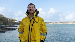 RNLI supporter Dermot O'Leary stands next to the sea wearing an RNLI all-weather lifeboat jacket. Photo: RNLI/Richard Adams